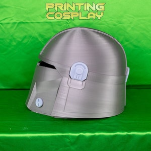 May include: A silver-colored Mandalorian helmet with white accents. The helmet has a visor and several detailed panels. The text "PRINTING COSPLAY" is displayed in a gold color at the top of the image. The helmet is on a green surface.
