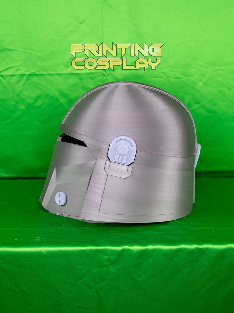 May include: A silver-colored Mandalorian helmet with white accents. The helmet has a visor and several detailed panels. The text "PRINTING COSPLAY" is displayed in a gold color at the top of the image. The helmet is on a green surface.