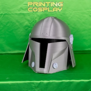 May include: A silver-colored Mandalorian helmet with a distinctive T-shaped visor and rounded features. The helmet has a textured surface and is set against a bright green background. The text "PRINTING COSPLAY" is displayed above the helmet.