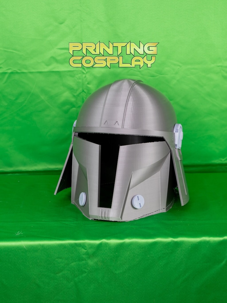 May include: A silver-colored Mandalorian helmet with a distinctive T-shaped visor and rounded features. The helmet has a textured surface and is set against a bright green background. The text "PRINTING COSPLAY" is displayed above the helmet.