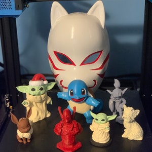 May include: A collection of figurines, including a white fox mask, a blue turtle, a green and white Yoda-like character, a brown and white Eevee, a red and black Deadpool-like character, a white and green Yoda-like character, and a white dog figurine.