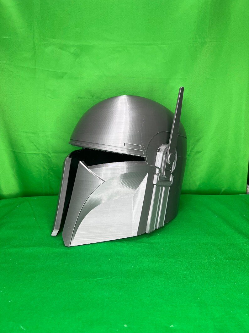 May include: A gray 3D printed helmet with a raised design and a small antenna on top. The helmet is angled to the left and has a textured surface.