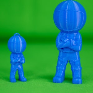 May include: Two blue 3D printed astronaut figurines, one larger than the other, standing with their arms crossed. The figurines are on a green background.