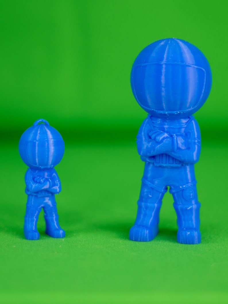 May include: Two blue 3D printed astronaut figurines, one larger than the other, standing with their arms crossed. The figurines are on a green background.