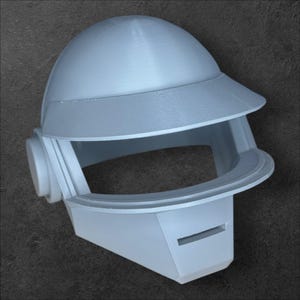 May include: A pale grey helmet featuring a visor and a curved top. The helmet has a rectangular lower section with a horizontal slit and a circular side component. The design is futuristic and geometric.