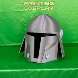 May include: A grey 3D printed helmet with a visor and side panels. The helmet has a textured surface and is designed to resemble a Mandalorian helmet.