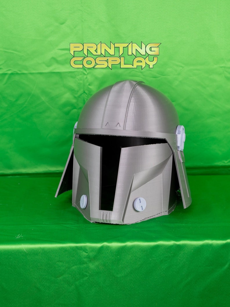 May include: A grey 3D printed helmet with a visor and side panels. The helmet has a textured surface and is designed to resemble a Mandalorian helmet.