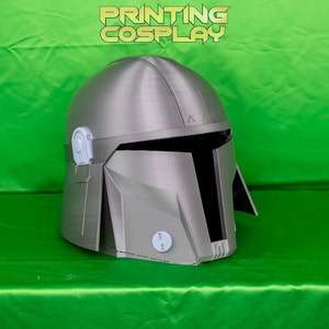 May include: A silver-colored Mandalorian helmet with a visor and a T-shaped opening. The helmet has a textured surface and is set against a green background. The text "PRINTING COSPLAY" is visible.