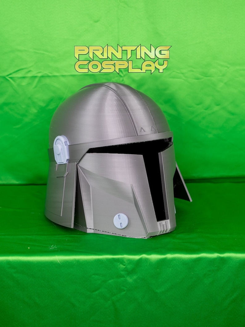 May include: A silver-colored Mandalorian helmet with a visor and a T-shaped opening. The helmet has a textured surface and is set against a green background. The text "PRINTING COSPLAY" is visible.