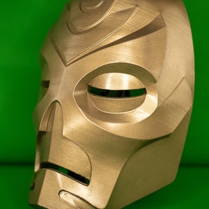 May include: A gold-coloured mask with a detailed design. The mask has a large, round eye opening and a smaller, rectangular mouth opening. The mask is made of a smooth, hard material.