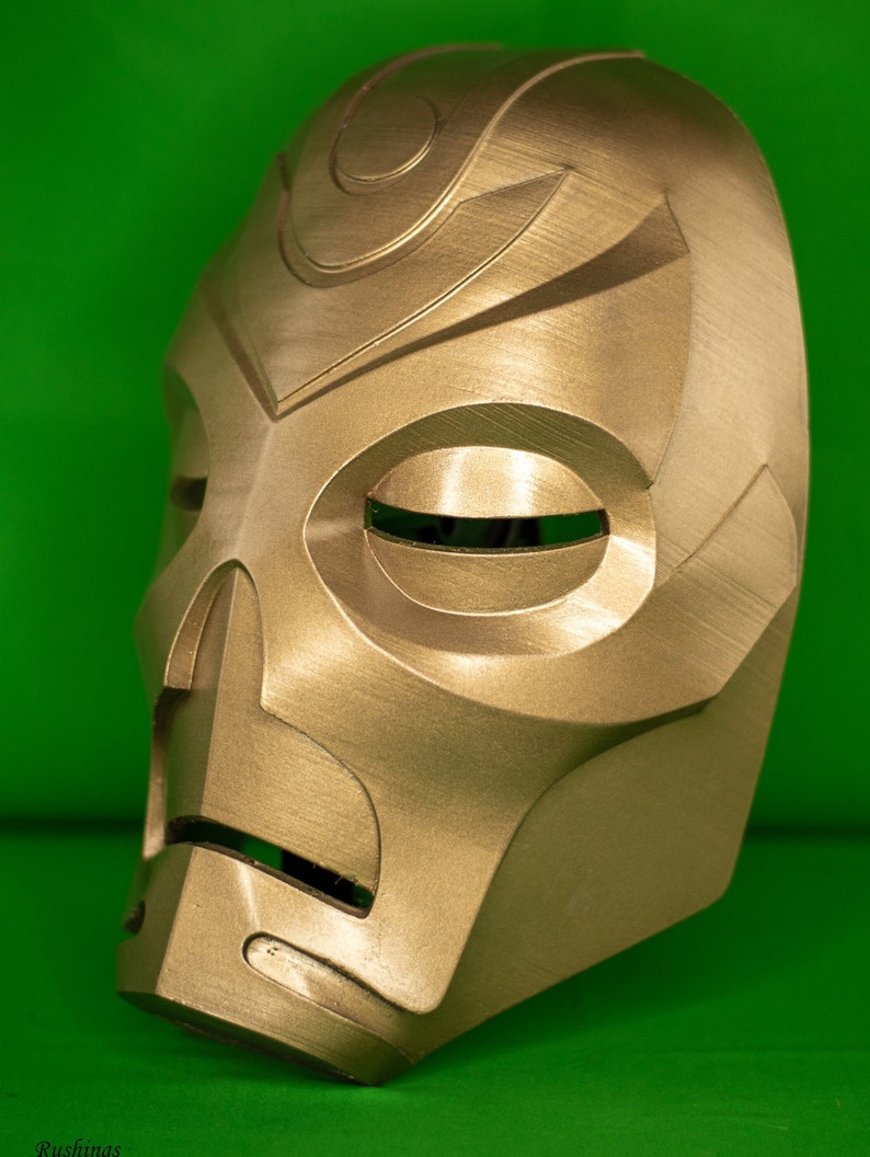May include: A gold-coloured mask with a detailed design. The mask has a large, round eye opening and a smaller, rectangular mouth opening. The mask is made of a smooth, hard material.