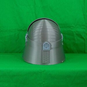 May include: A grey helmet with a textured surface and a small rectangular panel on the back. The helmet has a rounded top and a slightly pointed chin.