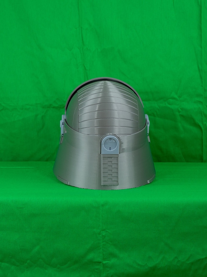 May include: A grey helmet with a textured surface and a small rectangular panel on the back. The helmet has a rounded top and a slightly pointed chin.
