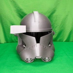 May include: A gray 3D printed helmet with a visor and a small rectangular attachment on the side. The helmet has a textured surface and a raised ridge on the top.