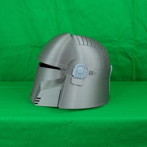 May include: A silver-coloured helmet with a textured surface and a raised design on the top. The helmet has a visor and a small, grey detail on the side.