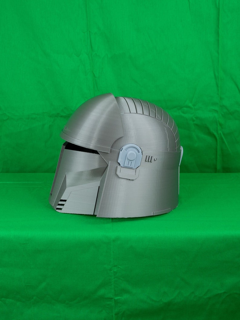May include: A silver-coloured helmet with a textured surface and a raised design on the top. The helmet has a visor and a small, grey detail on the side.
