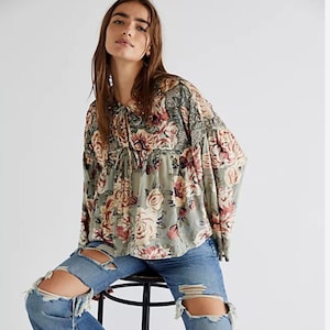 Puede incluir: Una blusa estampada con flores, mangas largas y detalle de lazo en el cuello. La blusa presenta un estampado de rosas rosas y rojas sobre un fondo verde. Combinada con vaqueros azules desgastados con rodillas rasgadas.