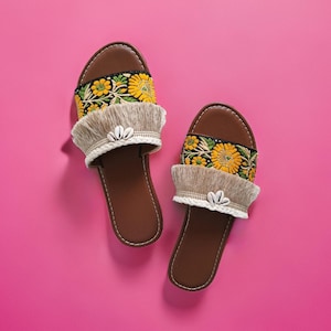 Könnte beinhalten: Ein Paar braune Ledersandalen mit Fransenbesatz und Blumenstickerei. Die Sandalen haben ein gelbes und grünes Blumenmuster und sind vor einem rosa Hintergrund platziert.