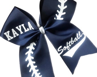 May include: A navy blue softball bow with white stitching details and the word "Softball" in a script font. The bow also features the name "Kayla" in white block letters. The bow is tied with a white ribbon.