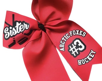 May include: A red cheerleading bow with the words "Sister" and "Arctic Foxes Hockey #3" printed on it. The bow features hockey stick and puck graphics.