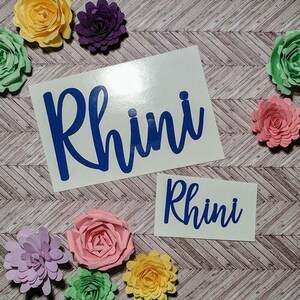 May include: Two white vinyl stickers with the name "Rhini" written in blue cursive lettering. The stickers are surrounded by colorful paper flowers.