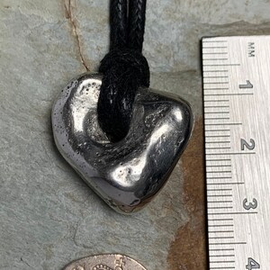 May include: A silver-colored pendant in a heart shape, suspended from a black cord. The pendant has a textured surface and a hole near the top. A ruler shows measurements in inches. A twenty pence coin is also visible.