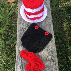 May include: A crocheted baby outfit in red, white, and black. The outfit includes a hat, a diaper cover, and a bow. The hat is a tall, red and white striped top hat. The diaper cover is black and has two red buttons. The bow is red and is tied in a bow.