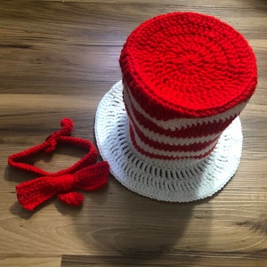 May include: A red and white crocheted hat with a matching bow tie. The hat has a wide brim and a tall, pointed crown. The bow tie is tied around the neck.
