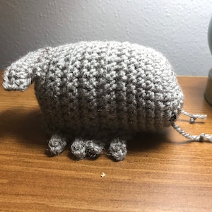 May include: A handmade, gray crocheted creature with a rounded body, small legs, and a curved trunk-like appendage. The amigurumi-style toy has small black eyes and two string antennae. The plushie is sitting on a wooden surface.