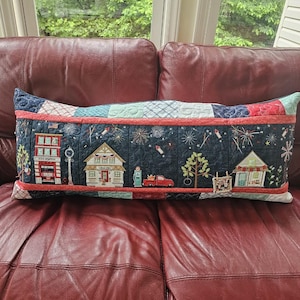 May include: A rectangular quilted throw pillow featuring a dark blue fabric panel with embroidered scenes of a fire station, a house, a red truck, and a small cottage. The design includes fireworks, trees, and a clothesline, framed by red piping and a patchwork border.
