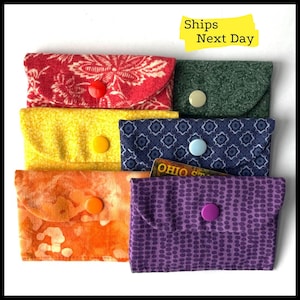 May include: Six colorful fabric pouches with snap closures. The pouches are stacked on top of each other and have different patterns. The top pouch is red with a floral pattern, the second pouch is yellow, the third pouch is blue with a geometric pattern, the fourth pouch is orange with a tie-dye pattern, the fifth pouch is purple with a polka dot pattern, and the sixth pouch is green with a paisley pattern.