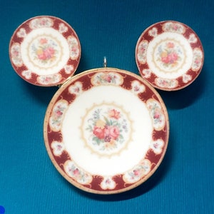 May include: A set of three decorative miniature plates. The plates have a white center with a floral design in the center. The rims are a deep red with a scalloped edge. The plates are arranged on a teal surface.