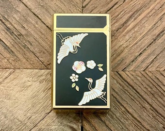 May include: A black and gold lighter featuring a white and gold design of cranes and flowers. The lighter is rectangular in shape and has a gold trim.
