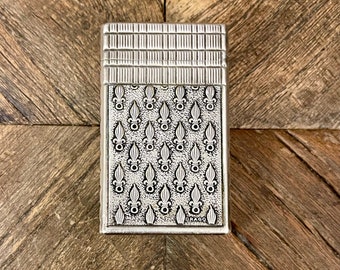 May include: Silver lighter with a repeating pattern of fleur-de-lis and the word "DRAGO" on the bottom.