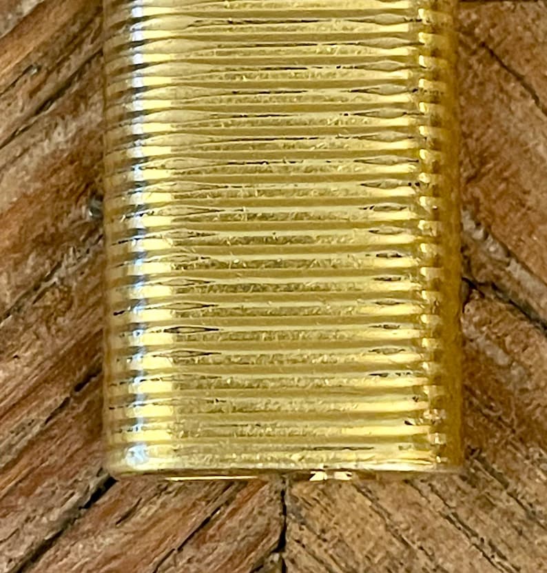 May include: Close-up of a gold-colored, rectangular lighter. The lighter's surface has a ribbed or lined texture, creating a vertical pattern. The gold tone appears consistent across the entire surface. The lighter's edges are clearly defined, and the overall impression is one of a high-quality, possibly luxury, item. The gold color is rich and lustrous, suggesting a metallic finish. The image focuses solely on the lighter, without showing any background or surrounding objects.