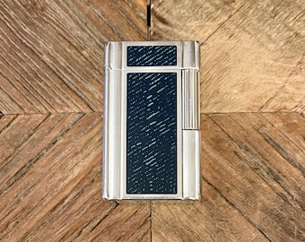 May include: A silver lighter with a dark green textured design on the front. The lighter has a silver trim around the edges.