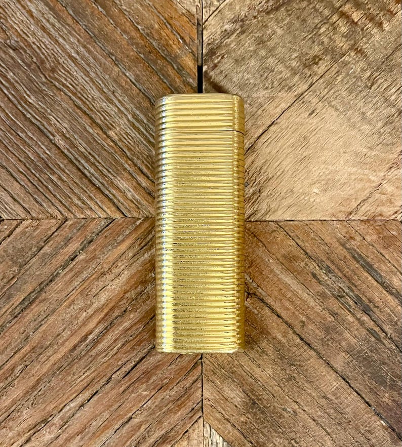 May include: A gold-tone, rectangular lighter with a ribbed texture. The lighter has a sleek, minimalist design and appears to be made of metal.  It is shown against a light brown wooden background. The lighter's surface is smooth and reflective, catching the light. The gold color is consistent throughout the lighter's body. This elegant accessory is perfect for adding a touch of luxury to any occasion.