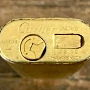 May include: Close-up of a gold Cartier Paris lighter. The lighter's top features the brand name and 'Swiss Made' markings.  The gold finish shows some wear.  The lighter's design is rectangular with rounded edges.  The lighter's mechanism is visible, showing a small, circular indentation for the flint.