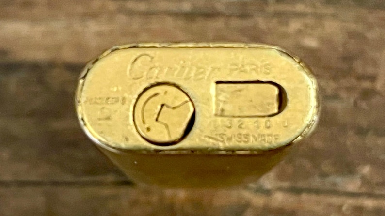 May include: Close-up of a gold Cartier Paris lighter. The lighter's top features the brand name and 'Swiss Made' markings.  The gold finish shows some wear.  The lighter's design is rectangular with rounded edges.  The lighter's mechanism is visible, showing a small, circular indentation for the flint.