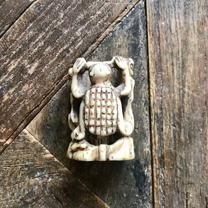 May include: A small, intricately carved ivory figurine of a turtle with its arms raised. The turtle is standing on a wooden surface.