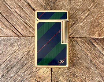 May include: A gold-coloured lighter featuring a navy, green, and red striped design. The rectangular lighter has rounded edges and a small logo. The lighter is placed on a wooden surface.