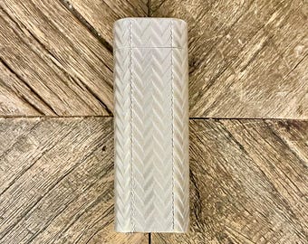 May include: A rectangular silver case with a textured chevron pattern. The case is closed and sits on a wooden surface with a diagonal pattern. The case has a hinged lid and a rectangular shape.