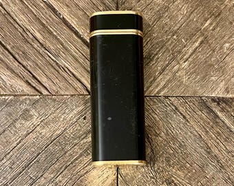 May include: A rectangular black lighter with gold-colored trim. The lighter has a glossy finish and is set against a wooden background. The top and bottom edges are accented with gold. The lighter is a classic design.