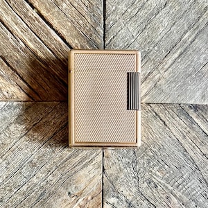 May include: A rectangular gold-toned metal lighter featuring a fine cross-hatched textured pattern across the front surface and a vertical ribbed rectangular accent panel on the right side, showcasing a metallic finish and a slim, flat design.