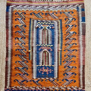 Könnte beinhalten: Ein rechteckiger Webteppich mit einem leuchtend orangefarbenen Feld und sich wiederholenden blau-weißen geometrischen Mustern. Die Mitte zeigt ein blaues Feld mit bogenförmigen architektonischen Mustern und rosa Akzenten, umrahmt von einer weiß-braunen Bordüre mit Fransen.