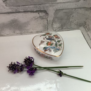May include: A white porcelain heart-shaped trinket box with a gold trim and a floral design featuring a peacock. The box is sitting on a white surface with lavender flowers.
