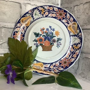 May include: A white ceramic plate with a blue and orange floral design. The plate has a scalloped edge and a raised rim. The floral design features a basket of flowers in the centre of the plate.