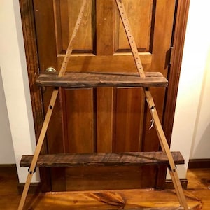 Può includere: Un'unità a scaffale in legno con tre ripiani, sostenuta da due gambe in legno inclinate con piedini in gomma nera. I ripiani sono realizzati in legno riciclato e hanno una finitura rustica.