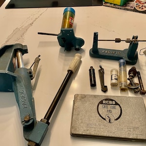 May include: A collection of reloading tools and supplies, including a blue Pacific reloading press, a blue Pacific case trimmer, a blue Pacific case lube pad, a white handle, a black case lube, and a gray RCBS case lube pad.