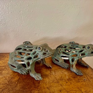 May include: Two bronze frog sculptures with openwork designs. The frogs have a verdigris finish, giving them a weathered appearance. They are positioned side-by-side, showcasing their detailed craftsmanship and unique aesthetic.